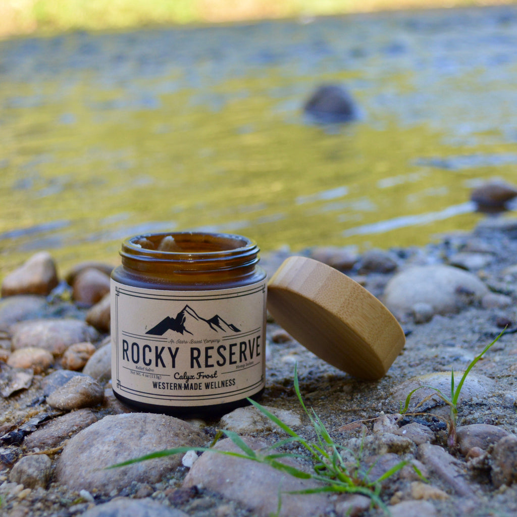 Candle jar labeled 'Rocky Reserve' on a rocky surface near a body of water.