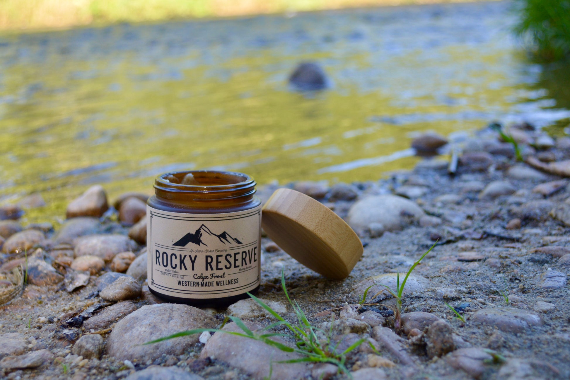 Candle jar labeled 'Rocky Reserve' on a rocky surface near a body of water.