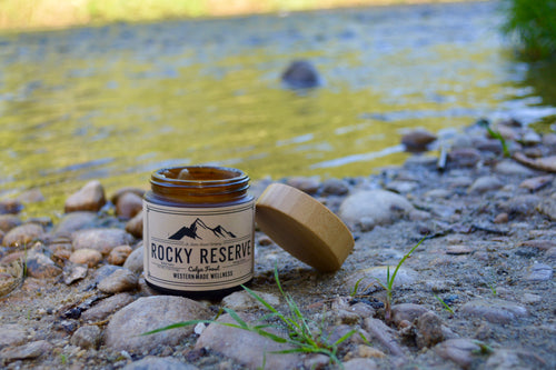 Candle jar labeled 'Rocky Reserve' on a rocky surface near a body of water.