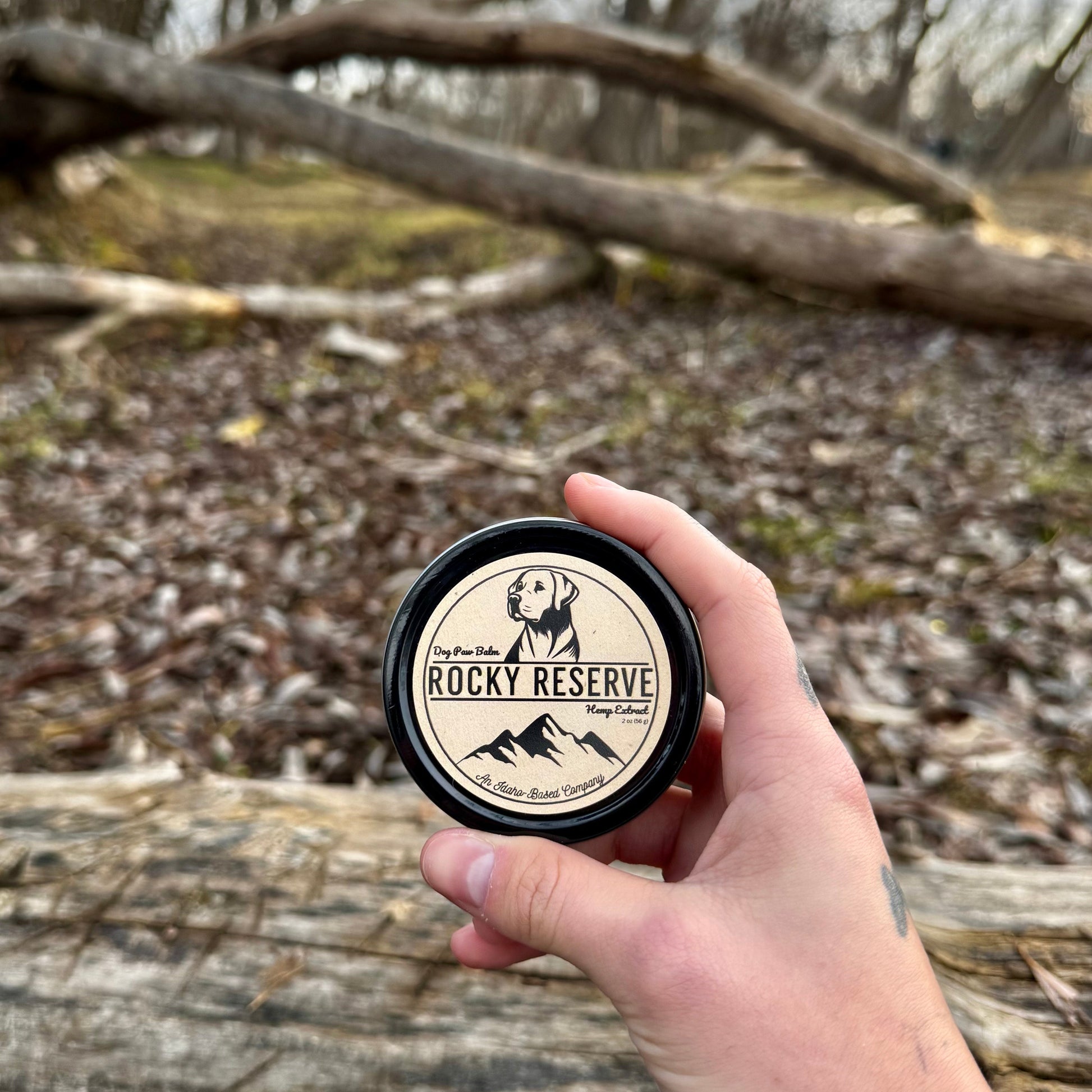 Hand holding a container labeled 'Rocky Reserve' against a natural background with trees and fallen branches.